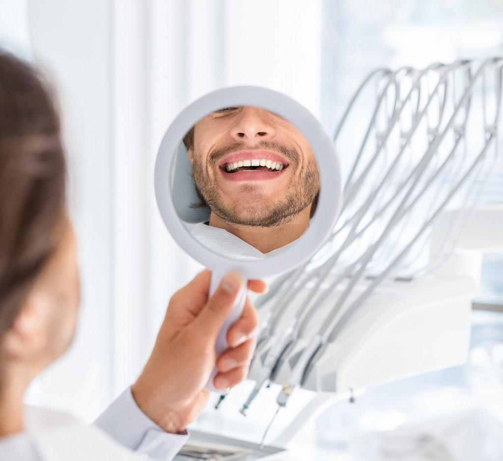 Handsome man looking at mirror and checking his beautiful smile, dental clinic interior, cropped